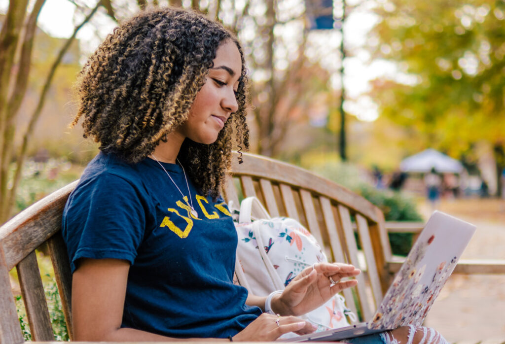 UNCG student seated on an outdoor bench with a laptop computer, with autumn trees in the background.
