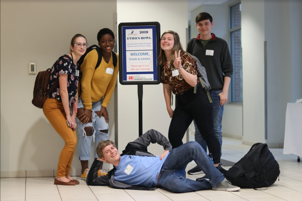 Five students smile and pose together around a sign reading "2019 Ethics Bowl"